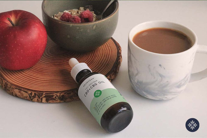 CBD + CBG Balance oil on a white table along with an apple, bowl of fruity oatmeal and coffee 