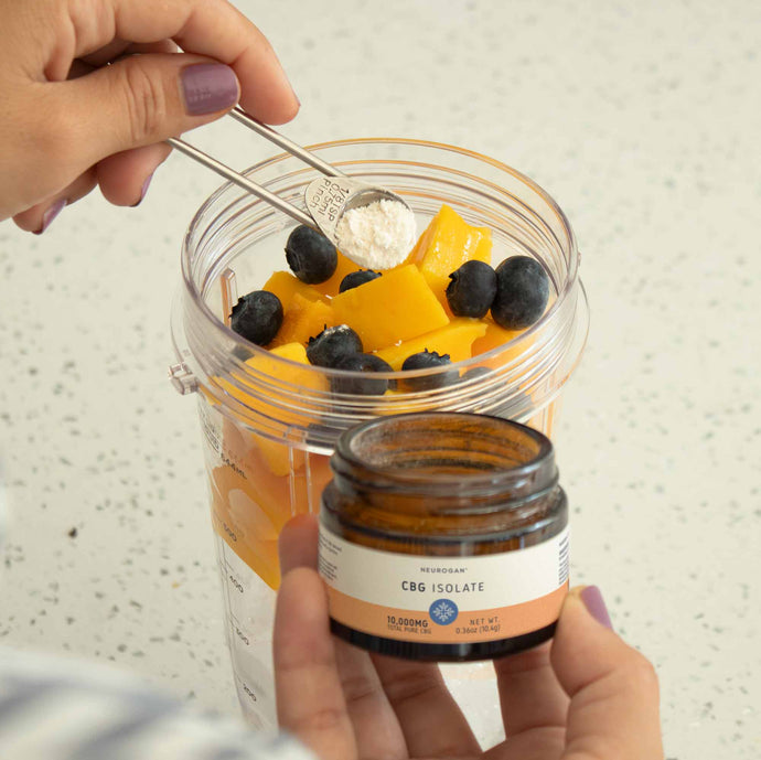 A scoop of CBG isolate powder being added to a blender filled with mango chunks and blueberries for a smoothie.