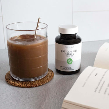 Bottle of CBD capsules beside a drink in a glass and an open book
