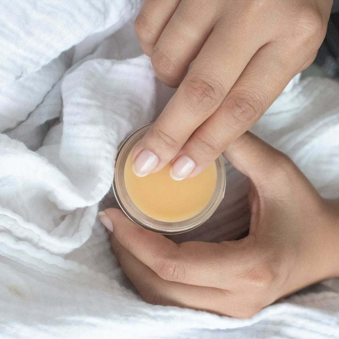 Woman scooping an amount of CBD salve from the jar placed on a white bed