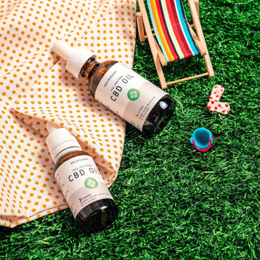 Two bottles of CBD Oil, laying on picnic blanket & grass lawn with beach accessories