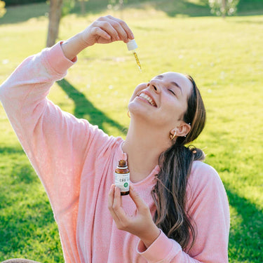 Woman smiling with eyes closed & sitting in a sunlit grass field, holding a dropper filled with CBD Oil