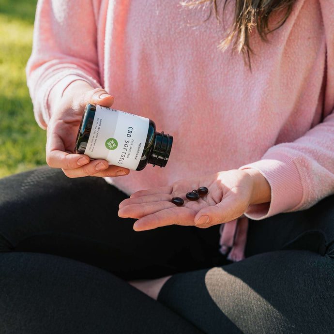 A woman sitting on a grass lawn, pouring three softgels into hand from a bottle of CBD Softgels