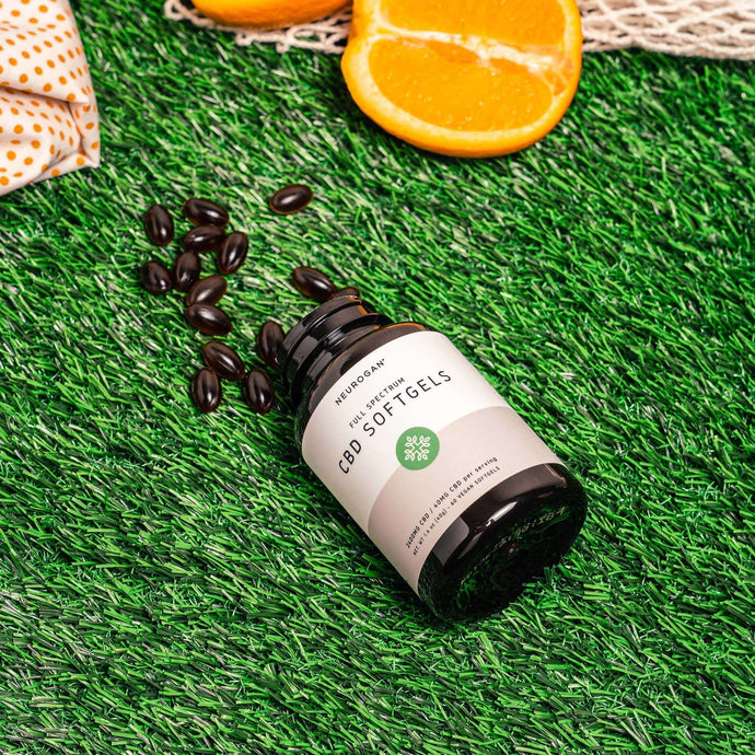 An open bottle of CBD Softgels, with 15 capsules spilled onto green grass lawn beside orange slices