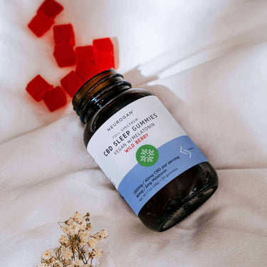 a bottle of CBD Sleep Gummies lays sideways on a bed with 10 red gummies poured onto white blanket