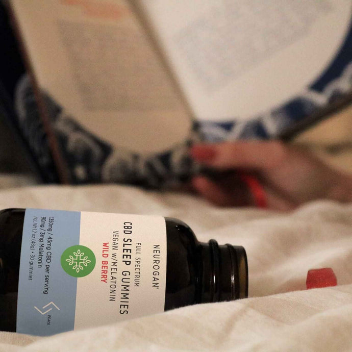 a bottle of CBD Sleep Gummies lays sideways on a bed with a red gummy poured onto white blanket